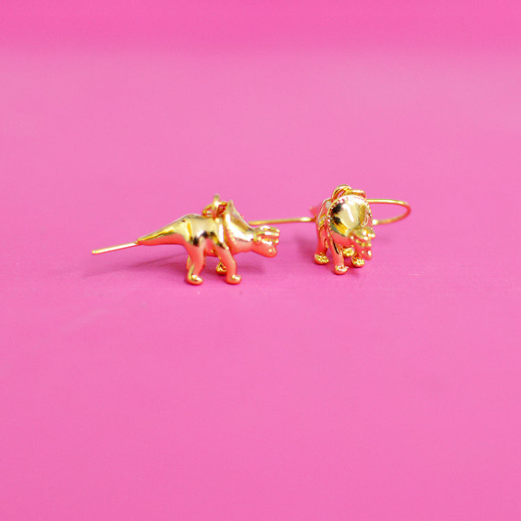 Gold plated triceratops shaped earrings on a pink background