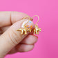 Gold plated triceratops dinosaur shaped dangle hook earrings held between fingers against a pink background