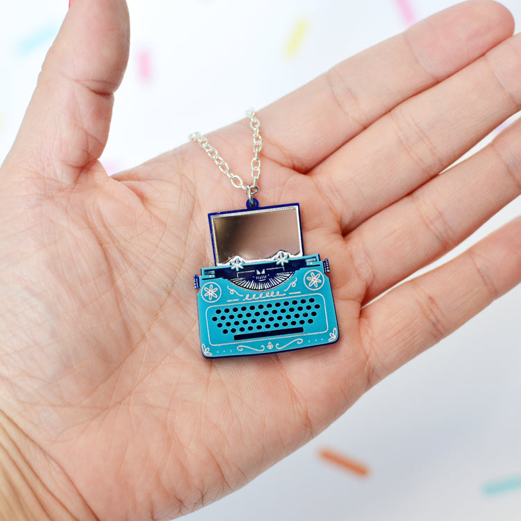 A miniature typewriter pendant necklace displayed on a person's palm. The pendant is bright teal blue with silver-toned metal details, including a silver chain, and is shaped like a vintage typewriter with paper coming out of the top that is made from mirrored material.