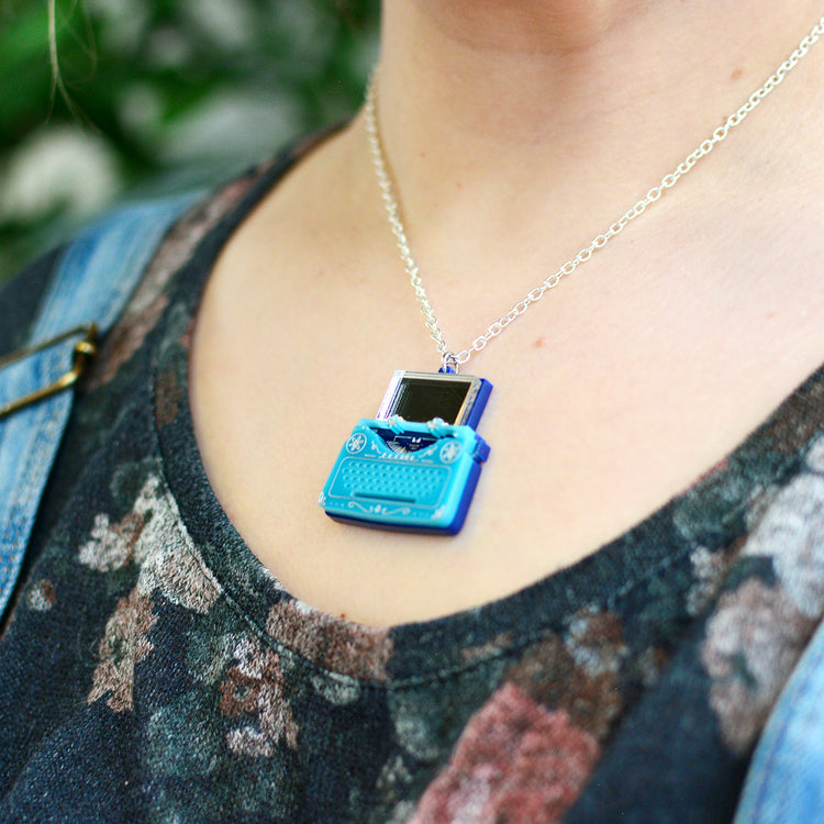 A close-up lifestyle shot of a person wearing a silver chain necklace with a miniature blue retro typewriter pendant