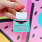 A hand with pink and yellow striped nail polish holds a turquoise typewriter-shaped earring with a blank white paper insert. The background is a colorful, abstract graphic pattern in pink, yellow, and blue.