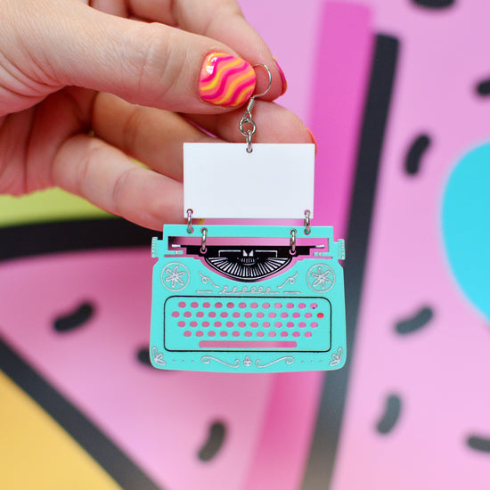 A hand with pink and yellow striped nail polish holds a turquoise typewriter-shaped earring with a blank white paper insert. The background is a colorful, abstract graphic pattern in pink, yellow, and blue.