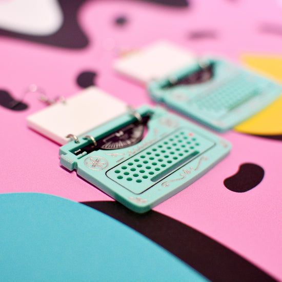 A close-up of mint green typewriter-shaped statement earrings with a white rectangular pendant, placed on a bright pink background with abstract black and blue shapes.