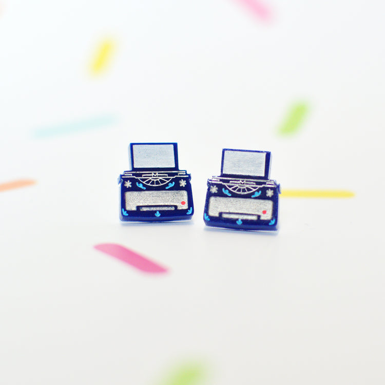 Typewriter shaped stud earrings on a white background with colourful shapes. The earrings are dark blue with silver coloured details.