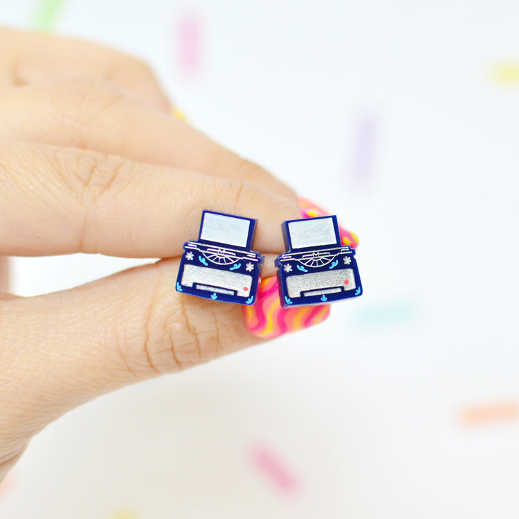 Typewriter shaped stud earrings held between fingers with a colorful blurred background