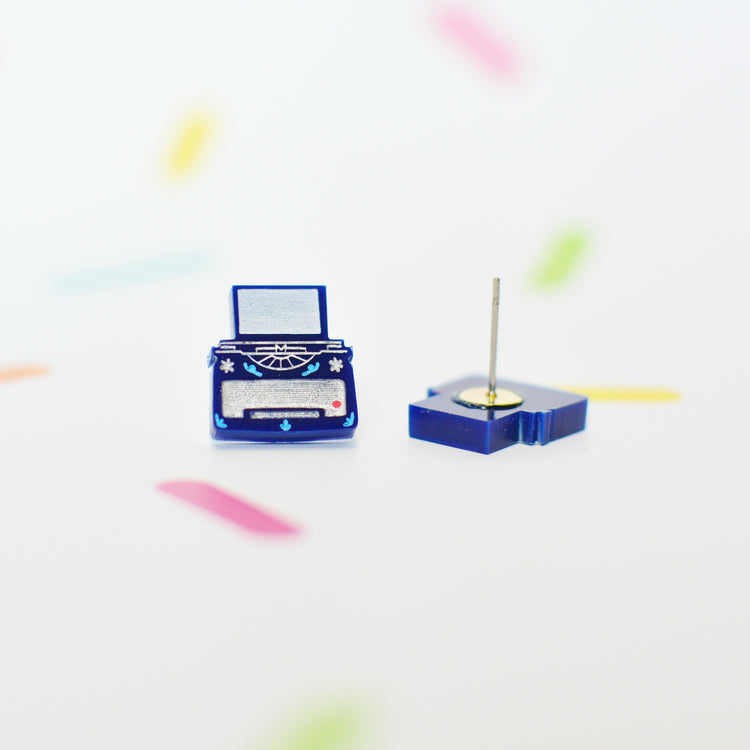 Small blue typewriter stud earrings on a white background with colorful abstract elements. You can see the whole of one stud earring and the back of the other which reveals the earring post.
